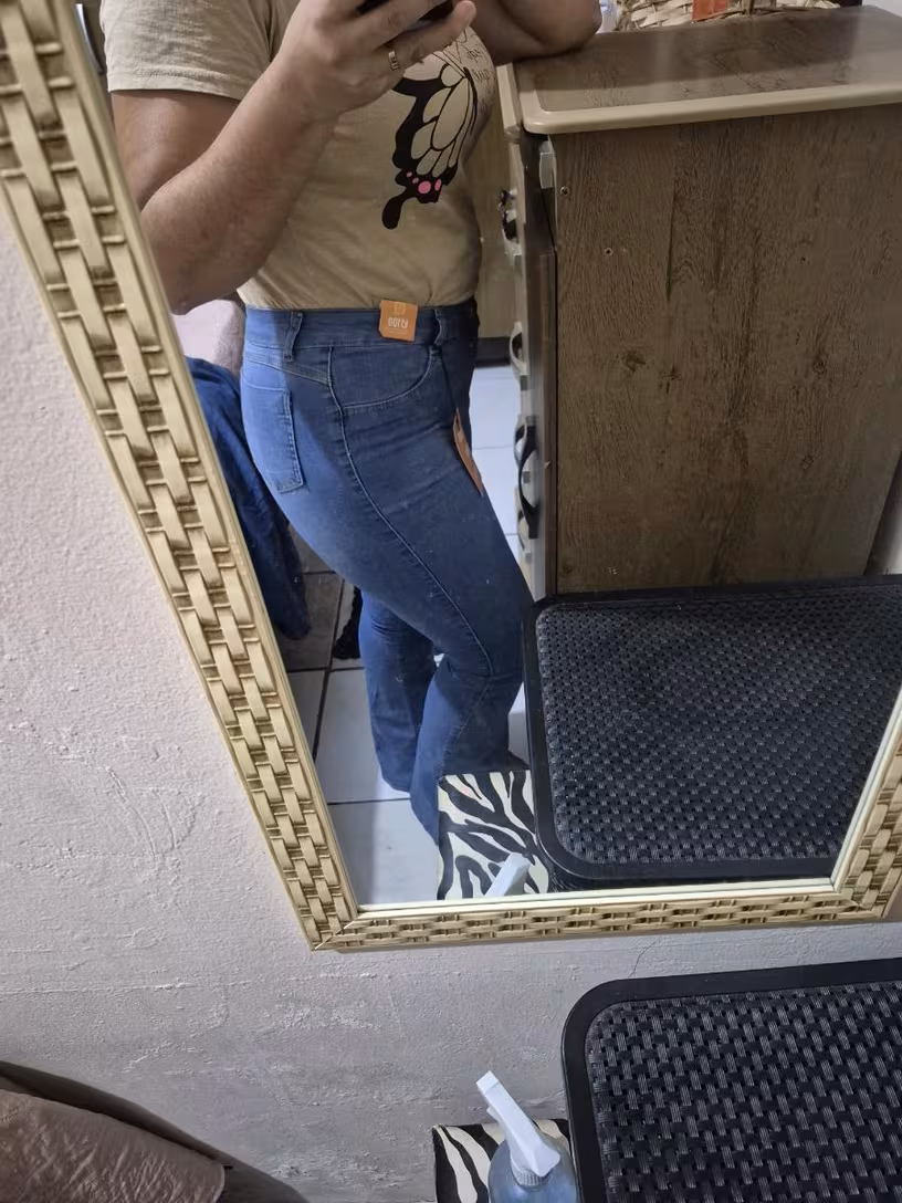 Reflexo no espelho de uma mulher vista de lado, vestindo calça jeans flare e camiseta, com etiqueta visível.