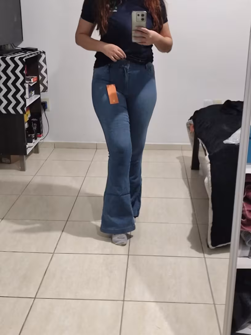 Mulher em frente a um espelho, vestindo calça jeans flare de lavagem média e camiseta escura, com etiqueta laranja pendurada.