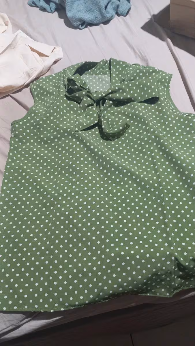 Regata plus size verde de bolinhas brancas com gola laço estendida sobre superfície de tecido claro