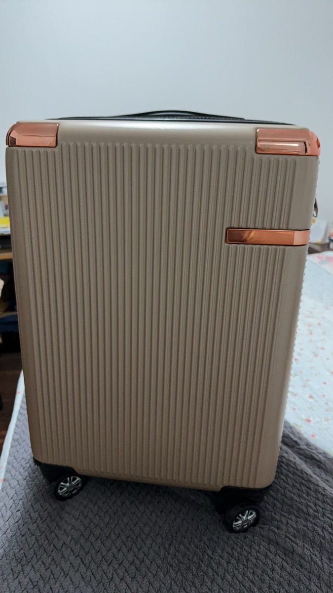 Mala de bordo dourada com detalhes em rosé gold e rodinhas pretas, em cima de uma cama.