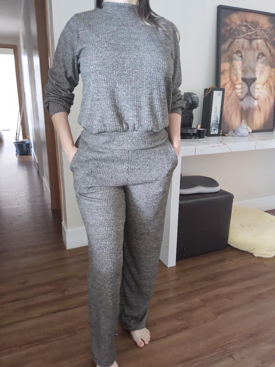 Modelo vestindo conjunto de moletinho canelado marrom com blusa de gola alta e calça reta, segurando uma bolsa preta em ambiente rústico.