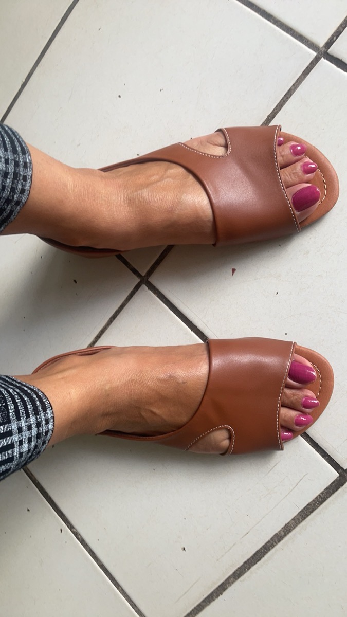 Pés de mulher calçando sandálias rasteirinhas em couro caramelo, com unhas pintadas de rosa, em um piso quadriculado.